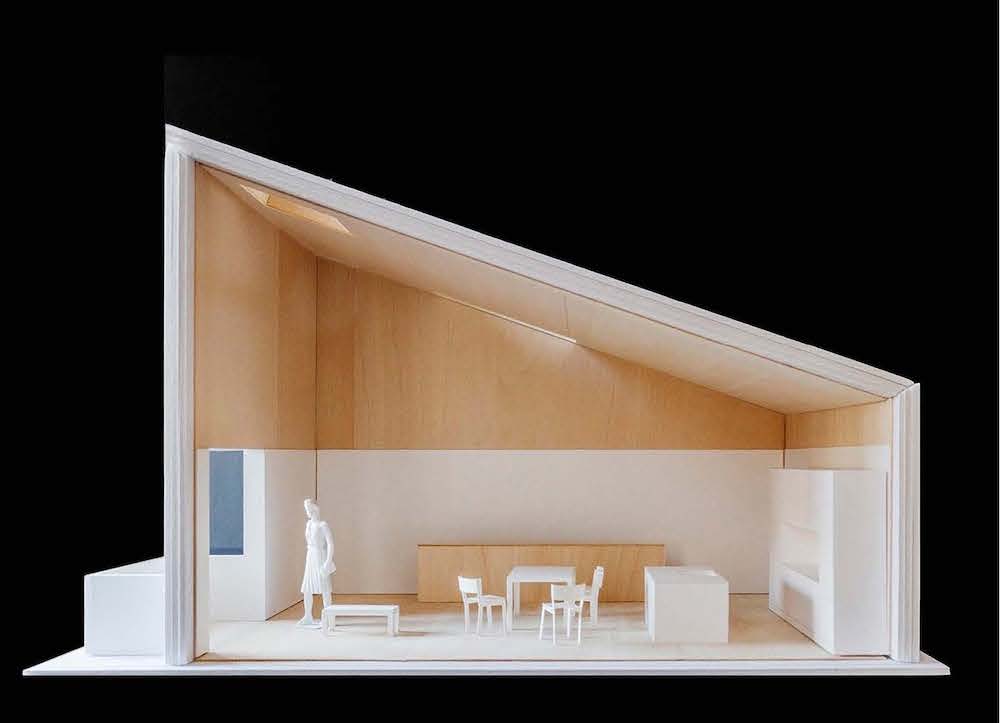 A side view of a wooden model of a single room with a slanted roof, with scale-model furniture and human figure