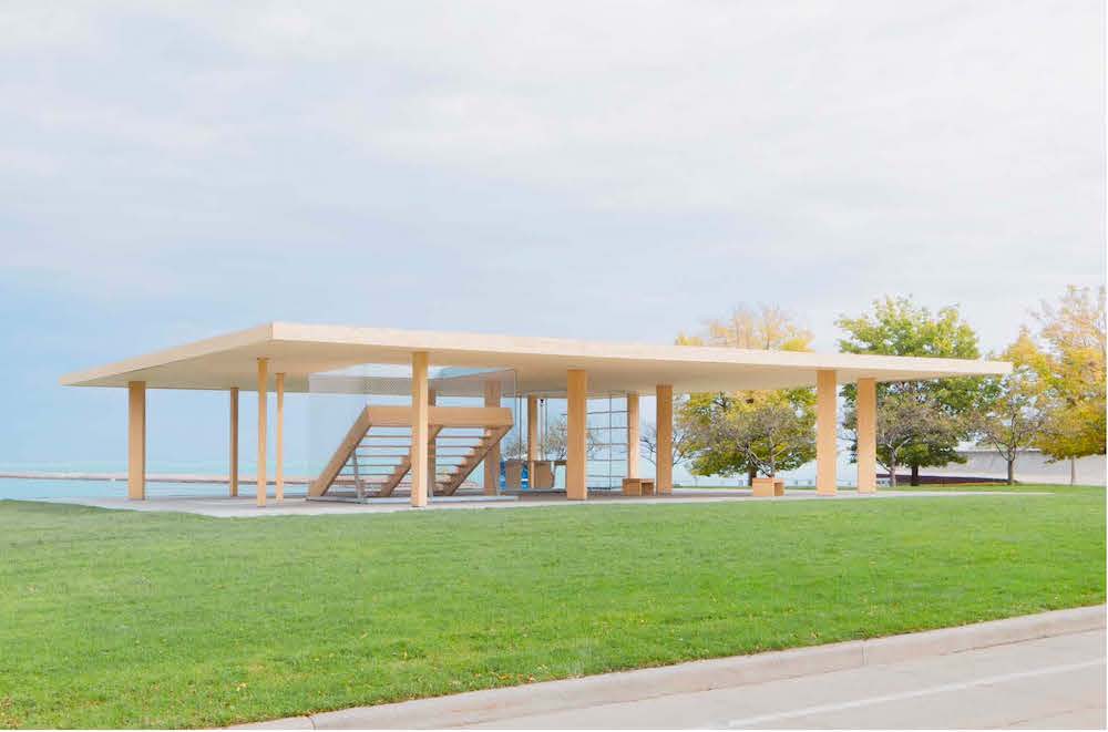 An open-side wooden structure with a prominent horizontal roof, support beams set amid a green lawn.