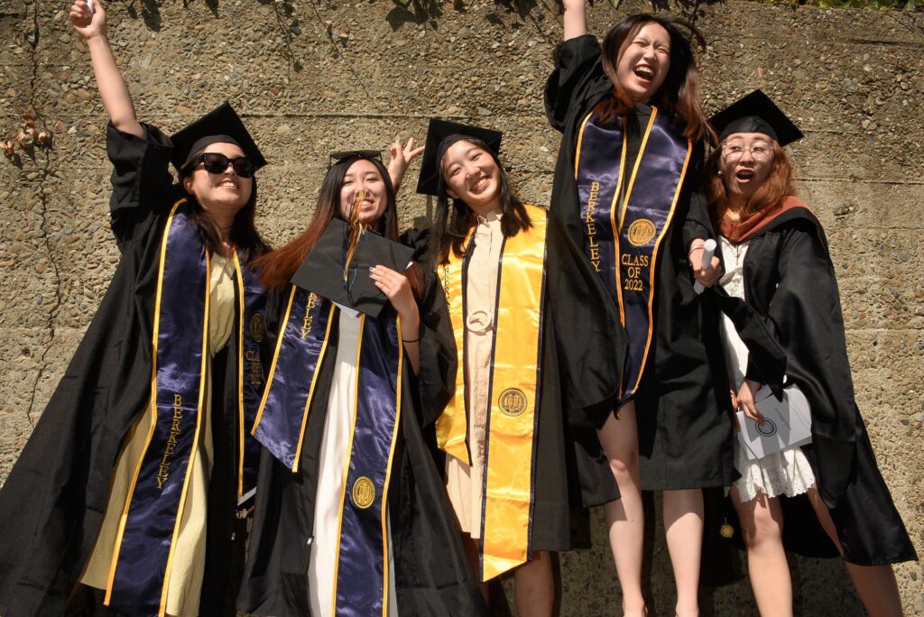 Five young women in graduation caps and gowns jump up in the air in celebration.