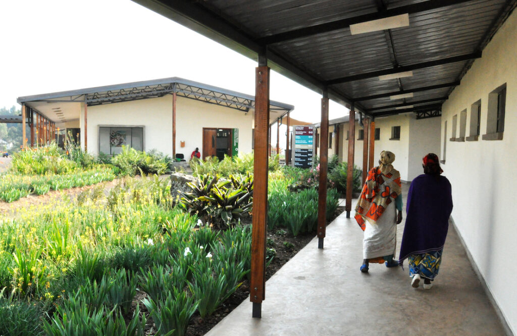 Two women in traditional African dress walk away from camera under covered walkway, with vegetation and a building in the background.