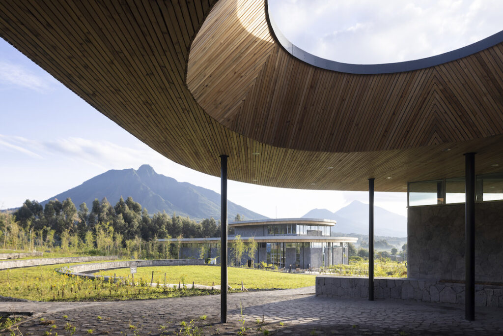 View of wooden structure open to the sky, with a glass facade building and mountains in background.