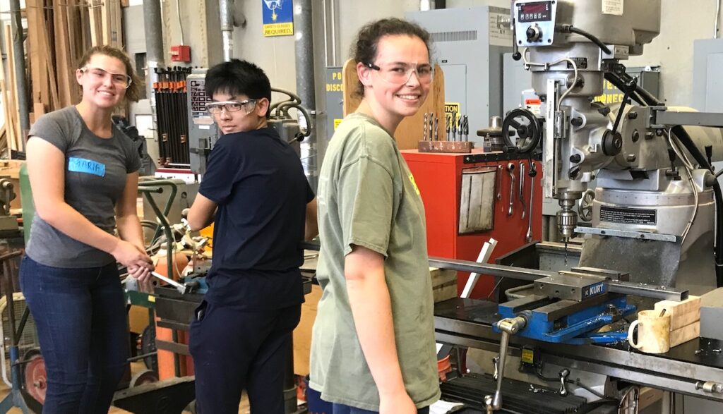 Teens wearing safety googles in a fabrication shop smiling at camera