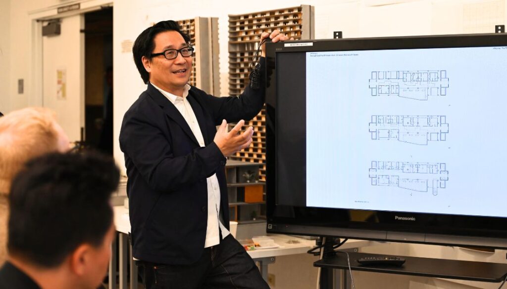 Professor teaching, gesturing to screen in front of audience