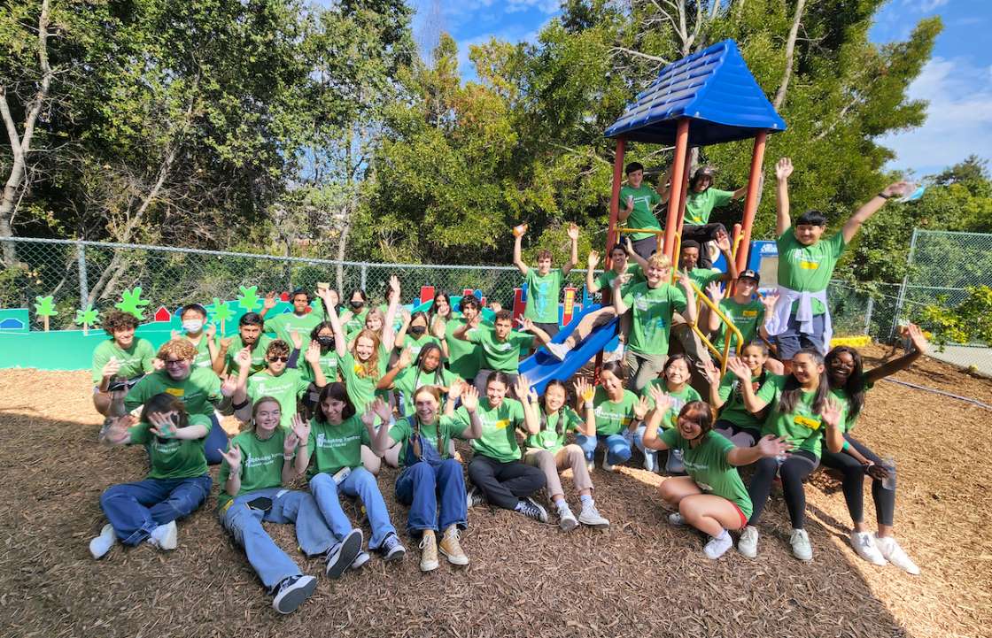 Students sitting on the ground around a playground structure posing for a group photo.